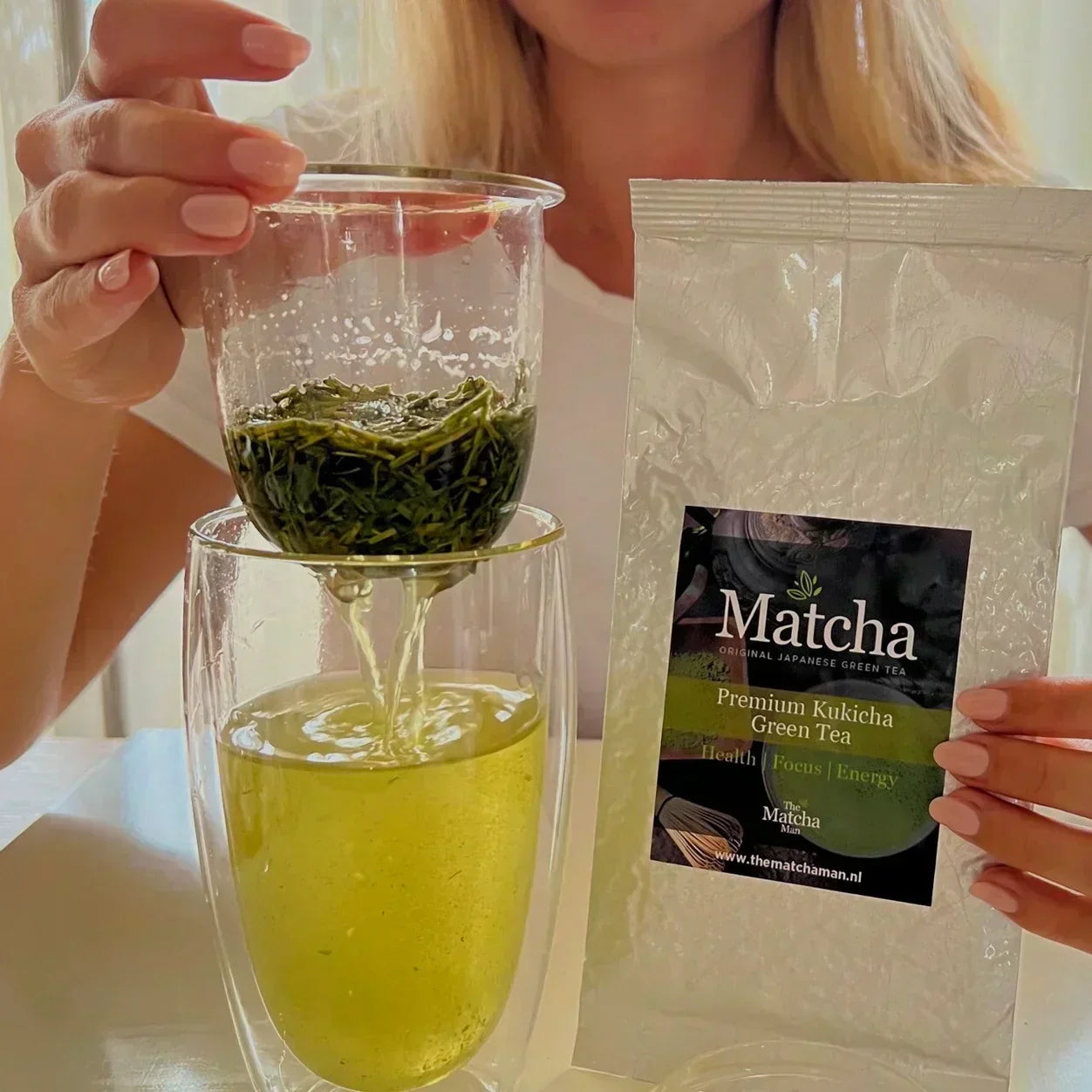 Dame toont zakje 'Premium Kukicha Green Tea'  van The Matcha Man en een glazen theefilter met losse thee boven een dubbelwandig glas gevuld met groene thee.